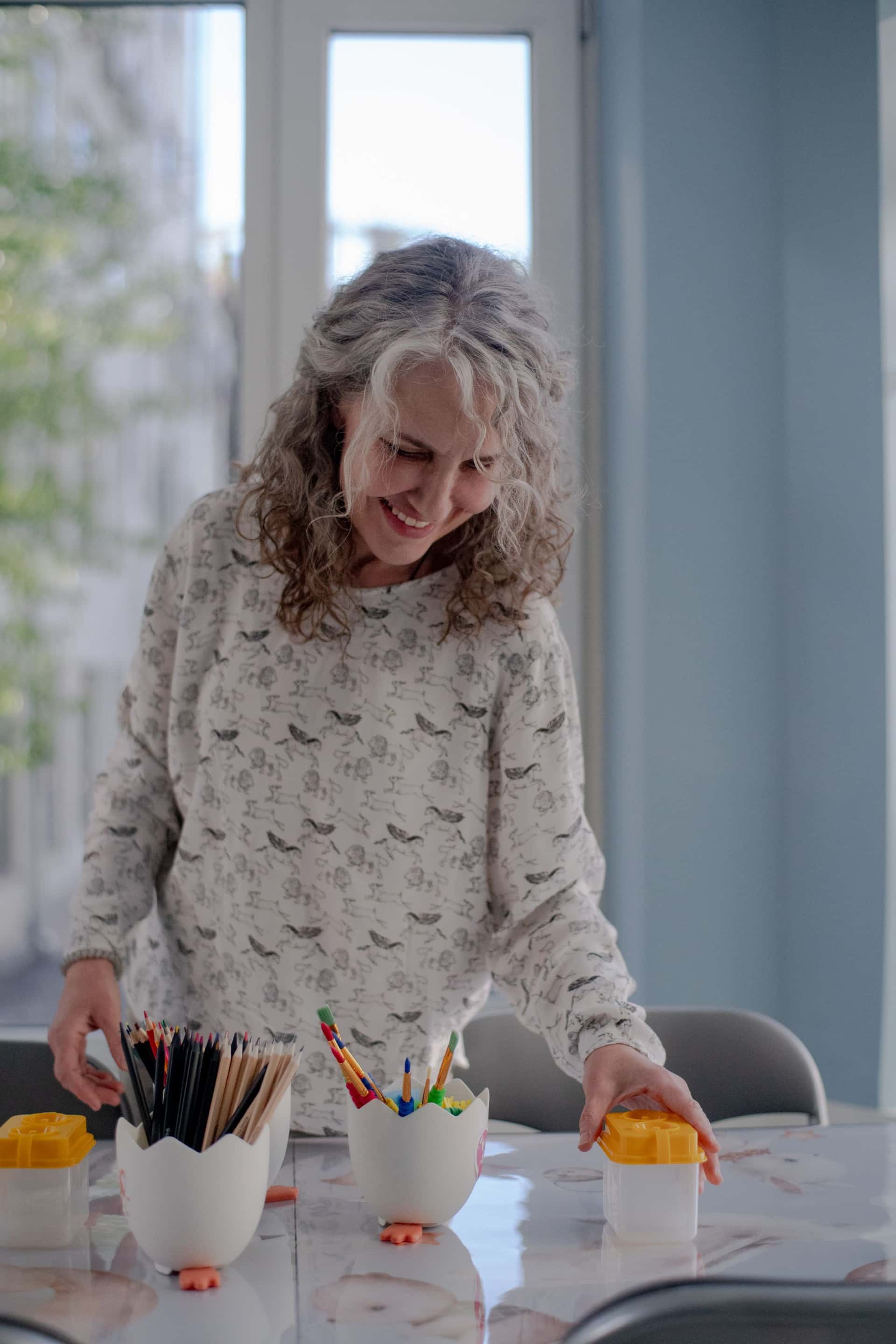 Женщина с кудрявыми волосами улыбается, раскладывая разноцветные ручки и карандаши по контейнерам на столе