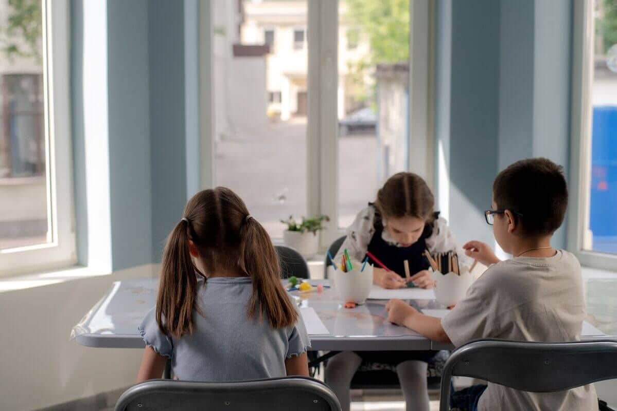 Трое детей сидят за столом с художественными принадлежностями и сосредоточенно рисуют в комнате