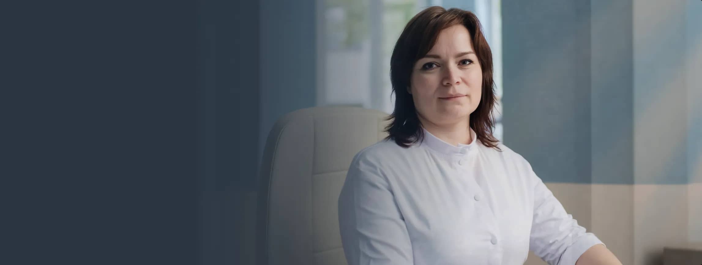 Женщина в белом халате сидит в светлой комнате, спокойно глядя в камеру