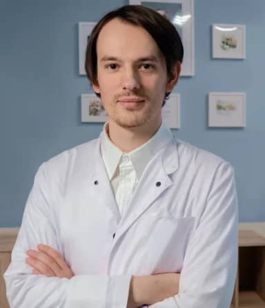 Человек в белом лабораторном халате уверенно стоит, скрестив руки, на фоне синей стены