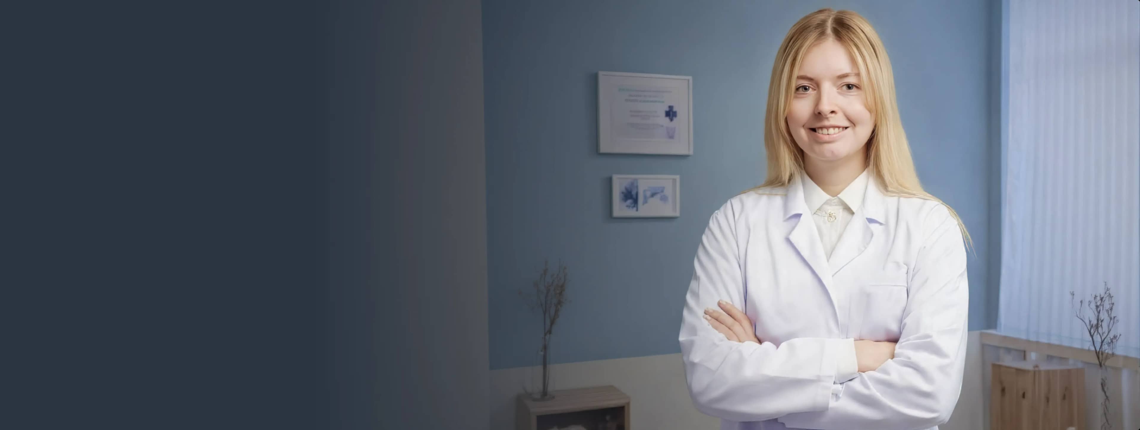 Улыбающаяся женщина в белом халате уверенно стоит, скрестив руки на груди, в светло-голубой комнате