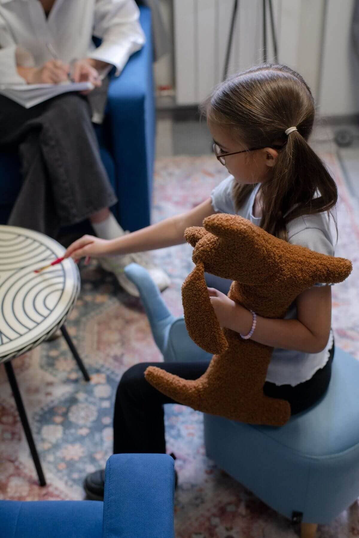 Молодая девушка в очках сидит на синем стуле, держа в руках коричневую мягкую игрушку