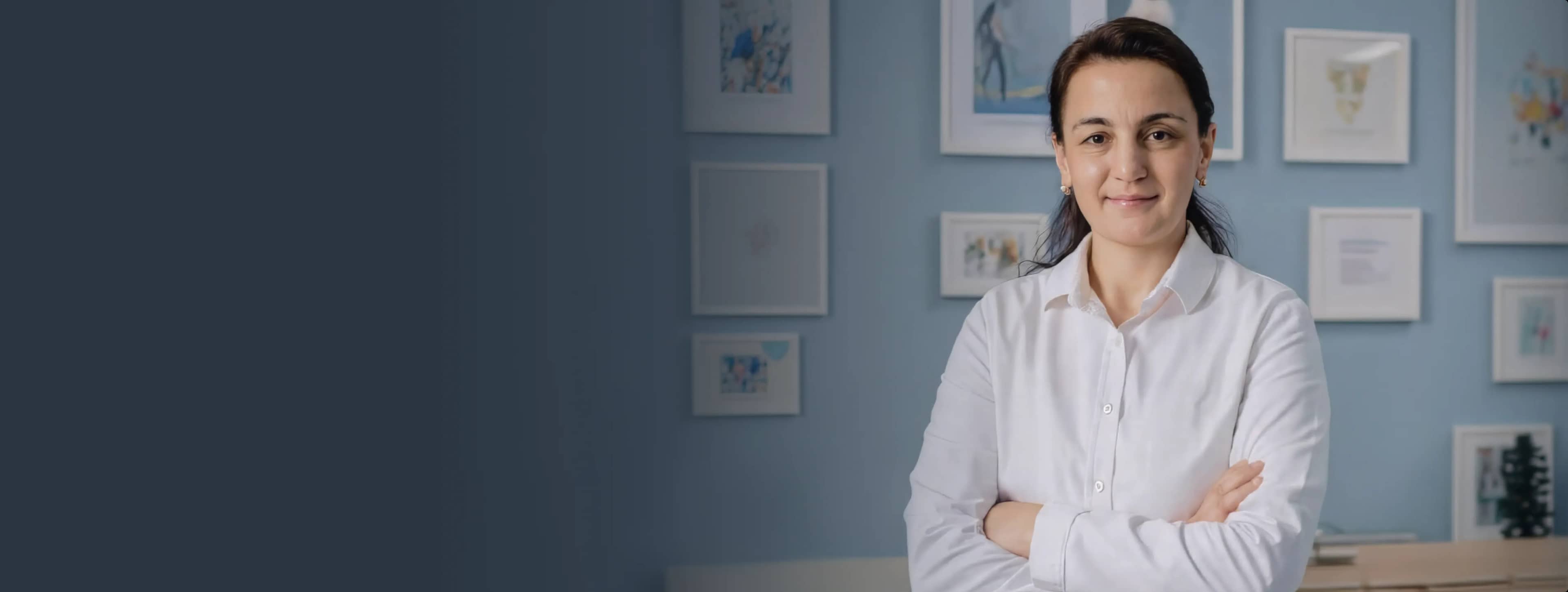 Женщина в белой рубашке уверенно стоит, скрестив руки на груди, и улыбается в офисе