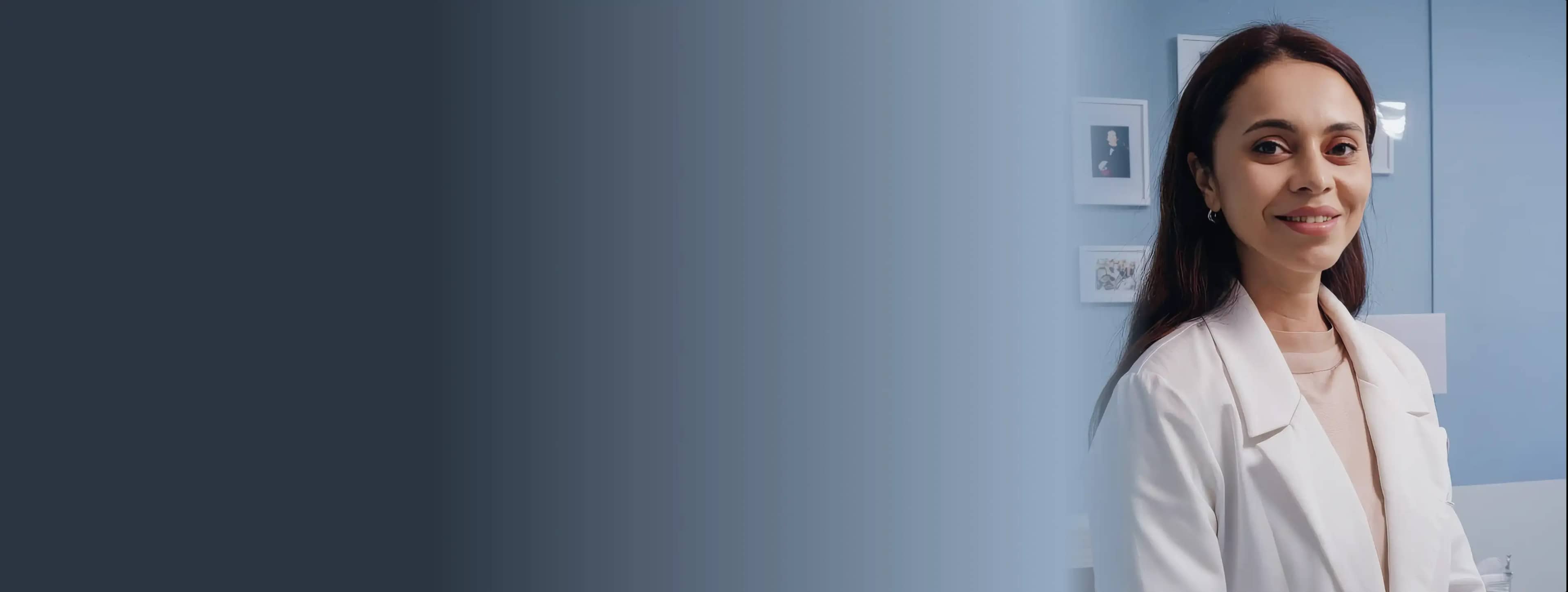 Женщина в белом халате, стоящая в светло-голубой комнате с фотографиями на стене и улыбается