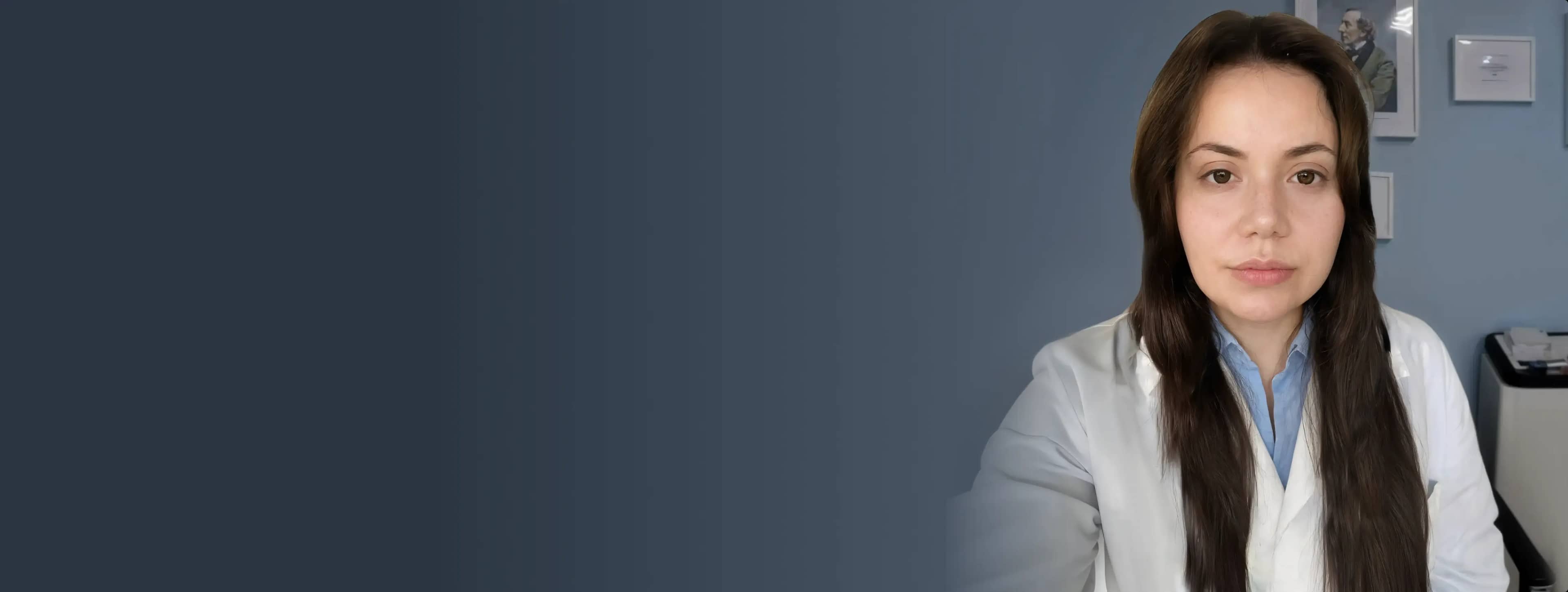 Женщина с длинными каштановыми волосами в белом халате и синей рубашке позирует в тускло освещённом офисе