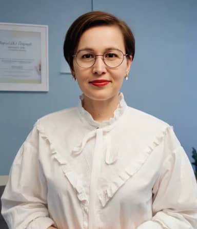 На синем фоне стоит человек в очках и белой блузке с рюшами