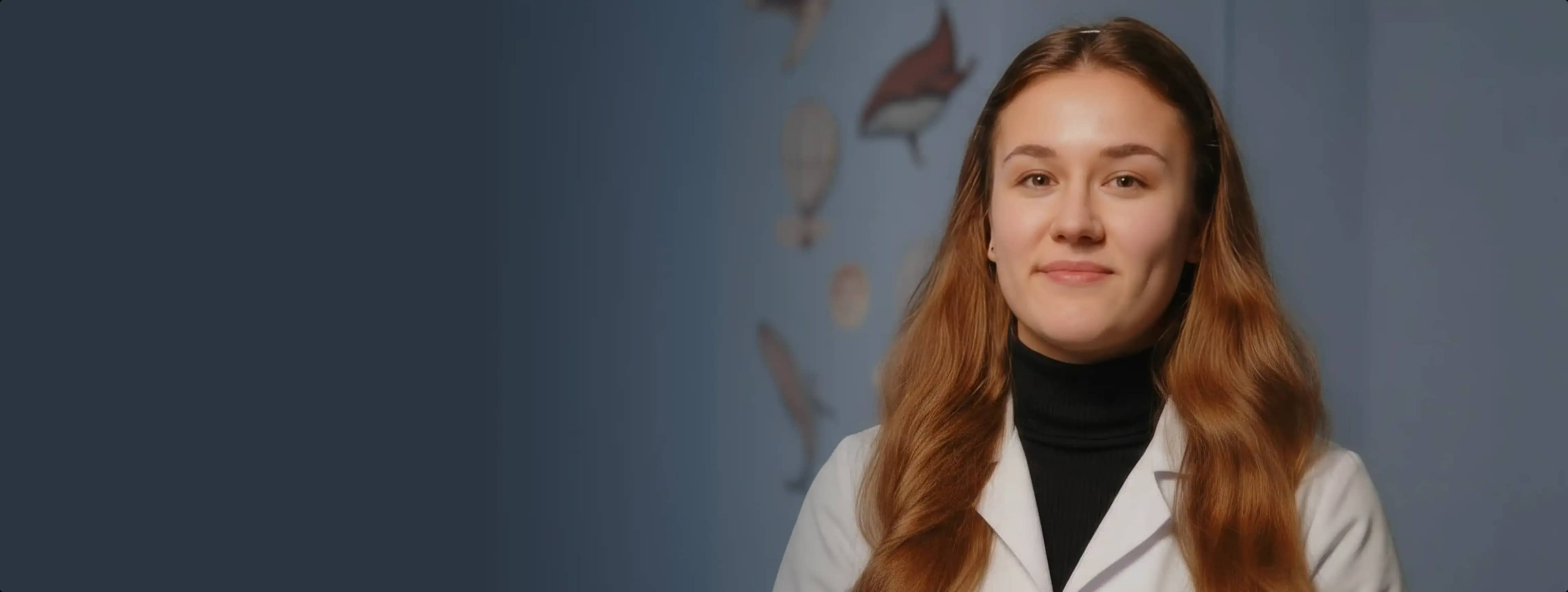 Женщина с длинными каштановыми волосами, одетая в белый лабораторный халат и черную водолазку