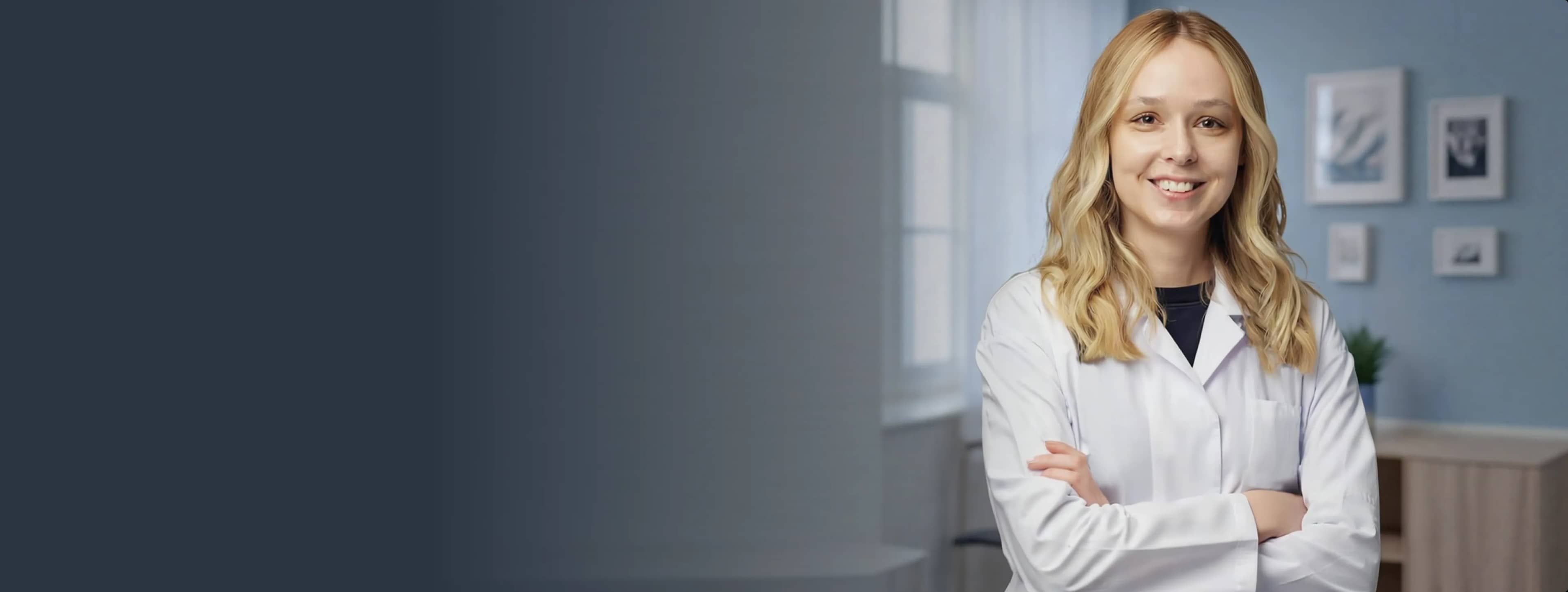 Улыбающаяся женщина в белом лабораторном халате стоит в мягко освещенной комнате с фотографиями в рамах на стене