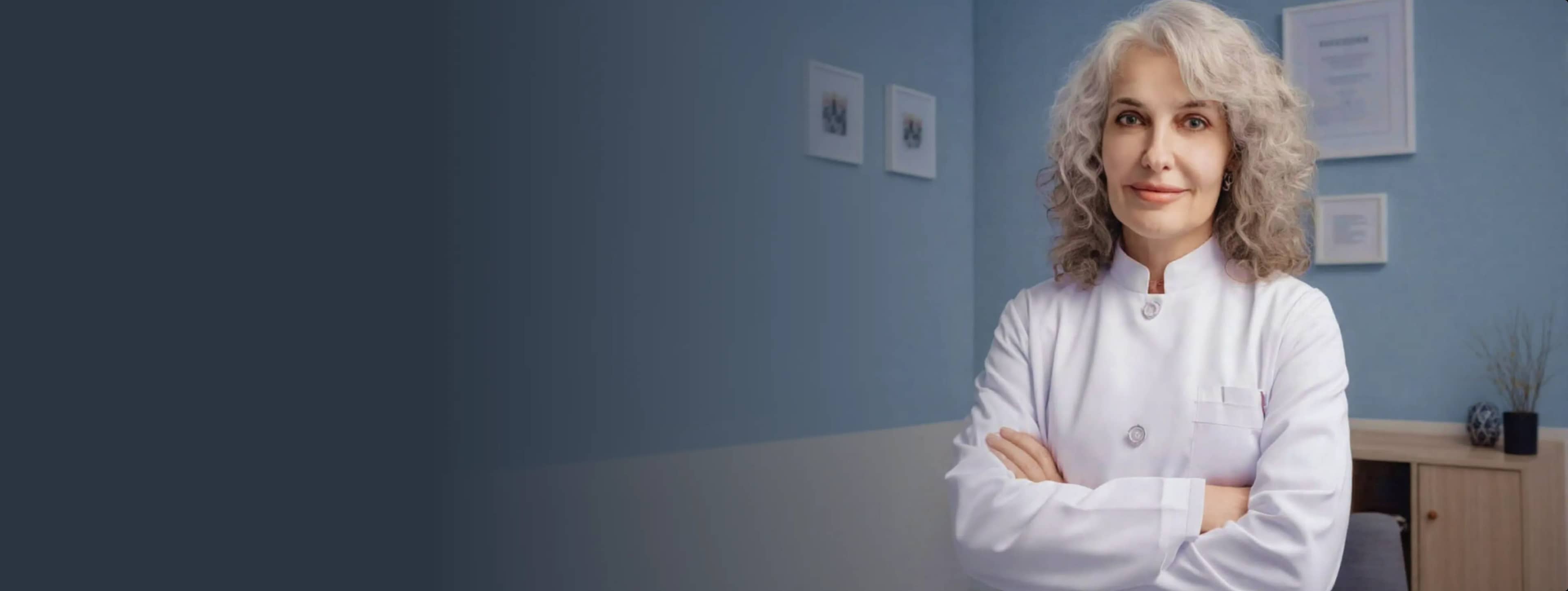 Женщина с вьющимися седыми волосами в белом халате, скрестив руки, на стене висят сертификаты