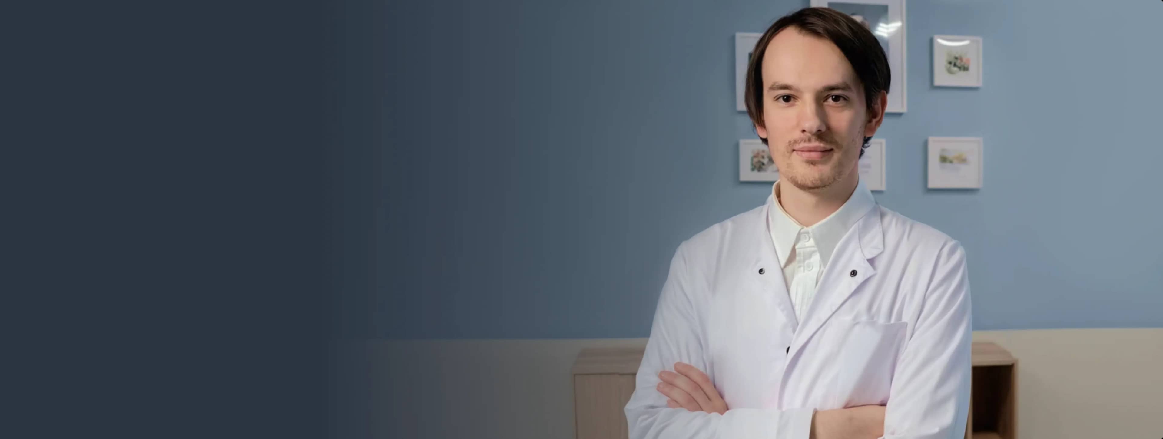 Мужчина в белом лабораторном халате стоит, скрестив руки, на фоне синей стены с фотографиями