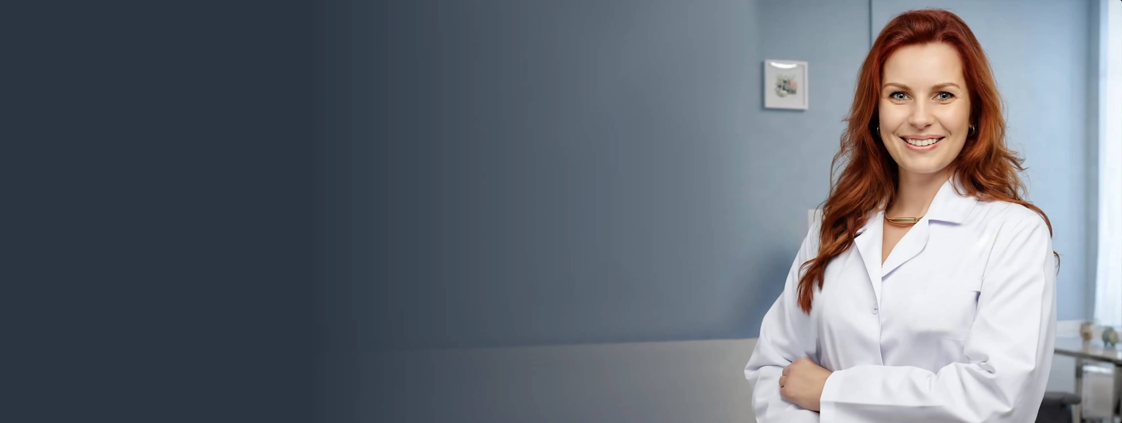 Улыбающаяся женщина-врач в белом медицинском халате на фоне светлой стены.