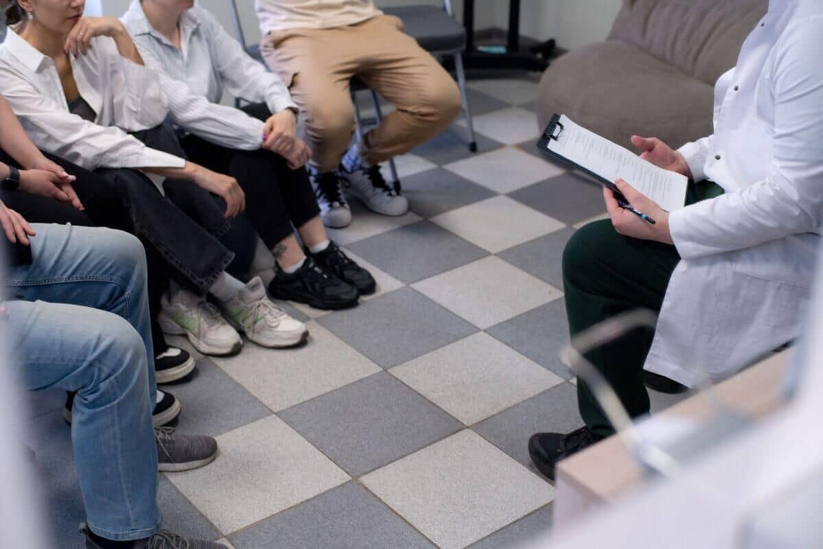 Группа людей сидит полукругом на стульях в комнате напротив человека в белом халате