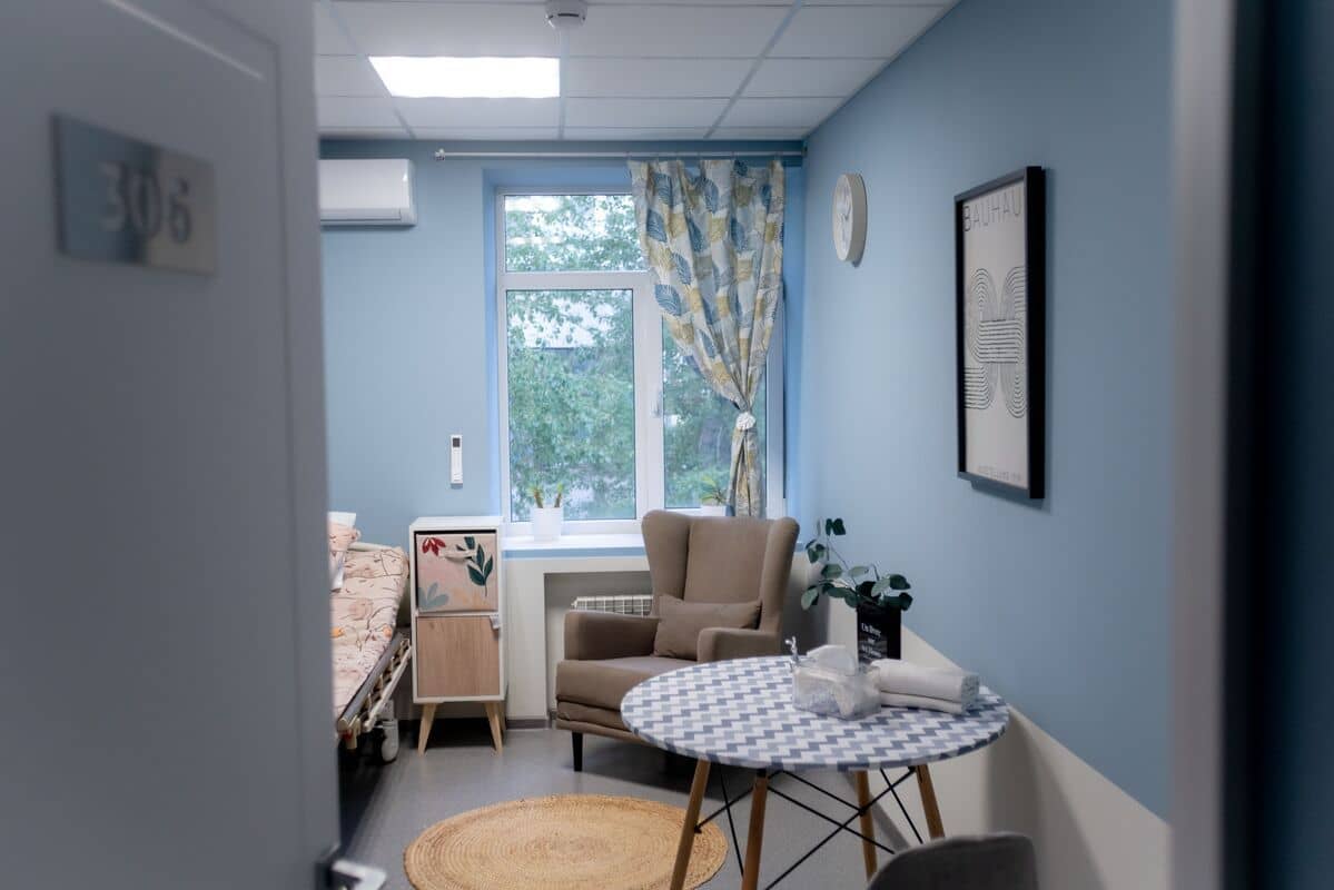 Больничная палата с бежевым креслом, круглым узорчатым столом и больничной кроватью