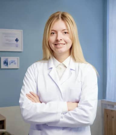 Улыбающийся человек с длинными светлыми волосами в белом лабораторном халате стоит